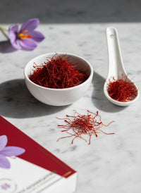 A pinch of Raihan Super Negin saffron threads beside a small bowl and porcelain spoon — a 1g bottle holds about 10 generous pinches